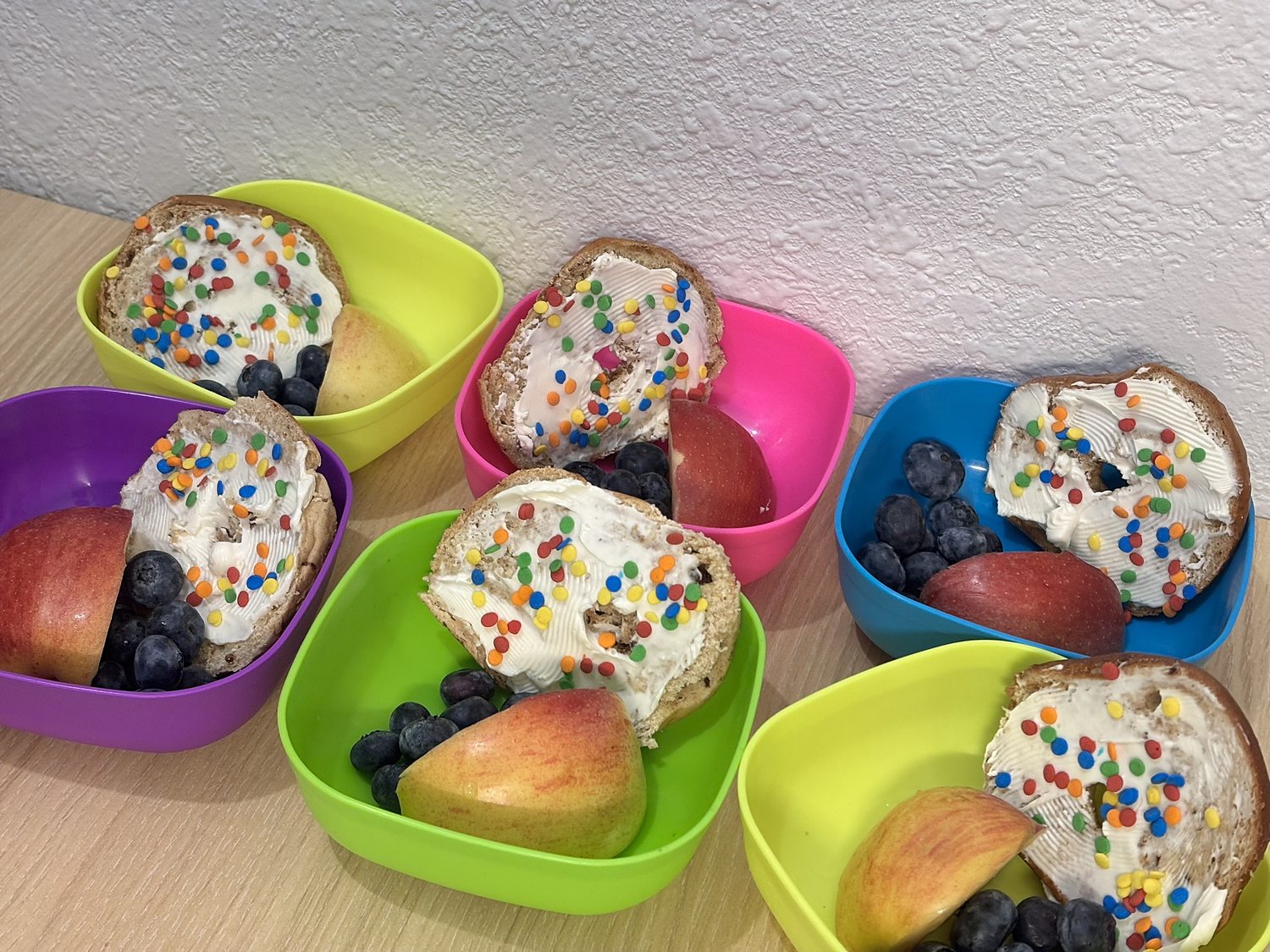 Bagels with cream cheese and sprinkles, apple slices and blueberries in colorful bowls